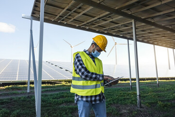 Young man working with digital tablet at renewable energy wind farm wearing safety mask for...