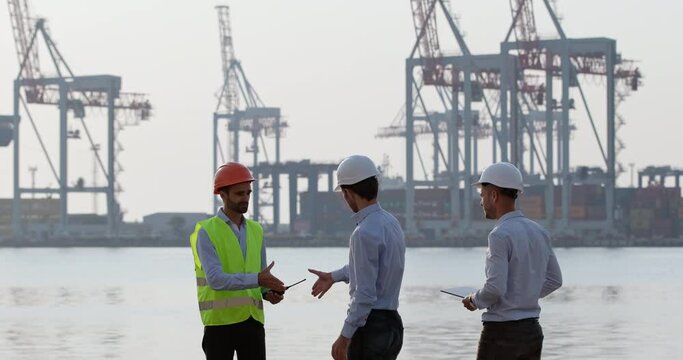 Men engineers and port worker wearing protective helmets discuss work issues concerning dockside cranes loading process on foggy morning