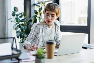 trendy businesswoman with tattooed body using calculator near laptop and blurred paper cup