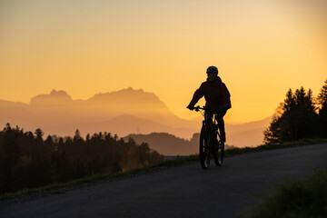 active woman riding her electric mountain bike at sunset in front of the awesome silhouette of Mount Saentis, Appenzell switzerland