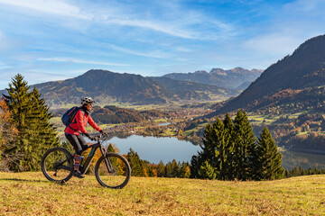 Obraz premium nice woman with electric mountain bike enjoying the view over lake Alpsee in atumnal atmosphere in the Allgaeu alps above Immenstadt, Bavarian Alps, 