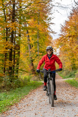 pretty senior woman ridin her electric bicycle in a colorful autumn forest with golden foliage
