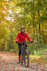 pretty senior woman ridin her electric bicycle in a colorful autumn forest with golden foliage
