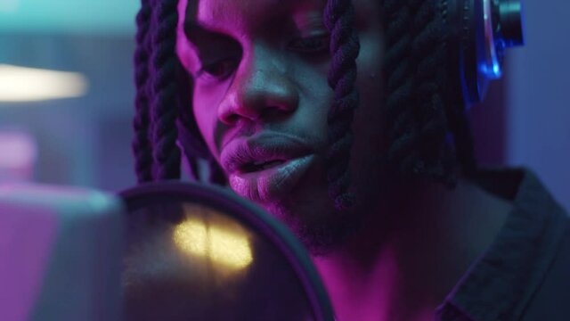 Close up shot of African American man with dreadlocks singing in microphone in recording studio with neon purple light