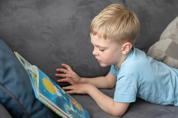 Cute little boy blonde reading a book lying on the couch