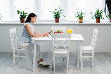 side view of pregnant woman reading book near orange juice and fresh apples on table in kitchen