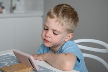Cute little boy blonde looks at the smartphone sitting at the kitchen at home. Concept online education, learning, games.