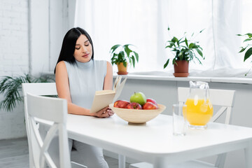 brunette woman reading book in kitchen near fresh apples and orange juice on table