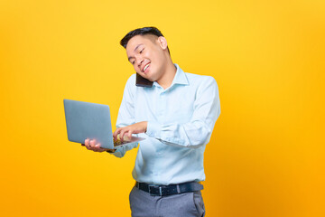 Young handsome businessman talking on a smartphone while holding a laptop on yellow background