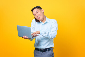 Young handsome businessman talking on a smartphone while holding a laptop on yellow background
