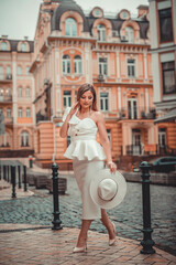 Close up fashionable woman portrait of young pretty fashionable girl posing in city in Europe, summer street fashion, holding retro hat. Fashion chic