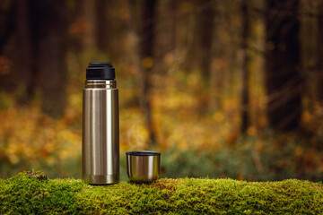Steel thermos with delicious hot tea on an old mossy tree in focus. The background of the autumn forest is blurred.
