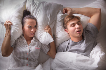 Woman looks at her boyfriend annoyed,holding pillow over her ears as he snores lying on his back...