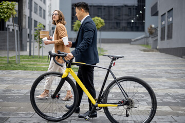 Fototapeta na wymiar Asian businessman and caucasian businesswoman walking, drinking coffee and talking in city. Concept of business cooperation. Idea of freelance and remote work. Man with bicycle. Young woman with food