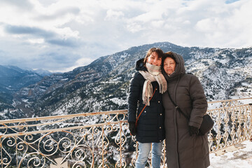 Winter portrait of hugging and smiling mother and daughter with splendid snow-covered mountains view in background, from Mega Spileon Monastery terrace at Vouraikos gorge. Winter vacation. Greece