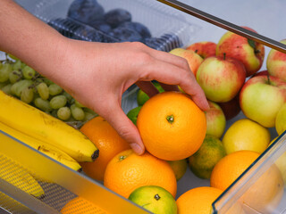 Woman taking orange from fridge drawer full of fruits.