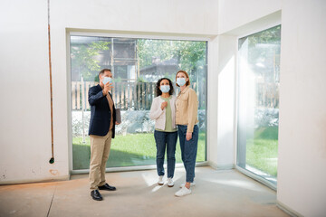 middle aged realtor in medical mask pointing with hand near interracial lesbian couple inside of new house
