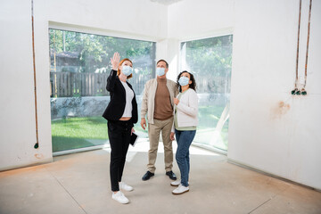 Fototapeta na wymiar redhead realtor in medical mask pointing with hand at new house while standing near interracial couple