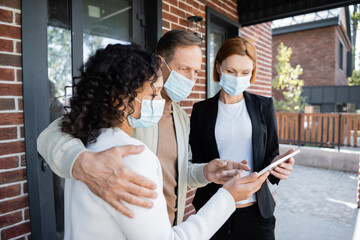 man in medical mask pointing at digital tablet near african american wife and realtor