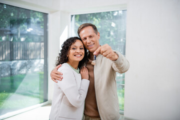 cheerful man holding blurred key and hugging african american wife in new house