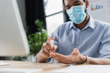 Fototapeta na wymiar Blurred african american businessman in medical mask spraying hand sanitizer near computer in office