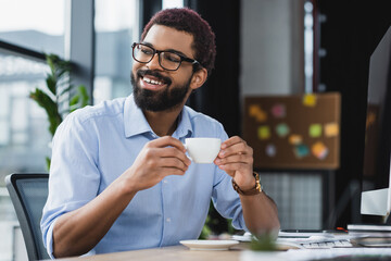Positive african american businessman in eyeglasses holding cup of coffee near computer in office