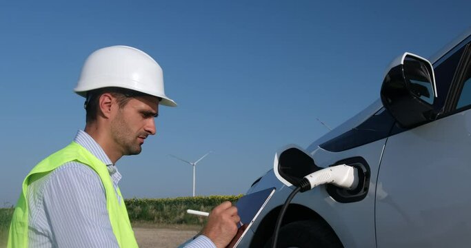 Wind turbines generate electricity for ev in sunflower field behind an engineer writing on tablet next to charging car
