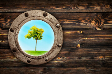 Close-up of an old rusty closed porthole window with  lonely tree standing on meadow view. Old rich wood grain texture background with knots.