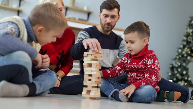 A married couple with children, sitting on the floor in a warm living room, playing with wooden cubes, wearing sweaters and having fun.
