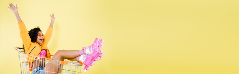 amazed african american young woman in pink roller skates sitting in shopping cart on yellow, banner