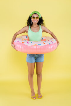 full length of joyful african american young woman standing with inflatable ring on yellow