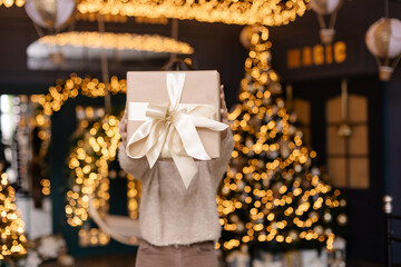 Fototapeta na wymiar Faceless man holding a box with a Christmas present in his hands. Gold ribbon gift wrapping. New year bokeh background copyspace place for text. Festive interior