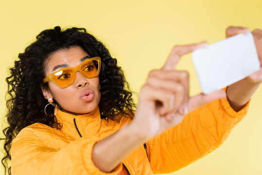 curly african american young woman in sunglasses pouting lips and taking selfie isolated on yellow