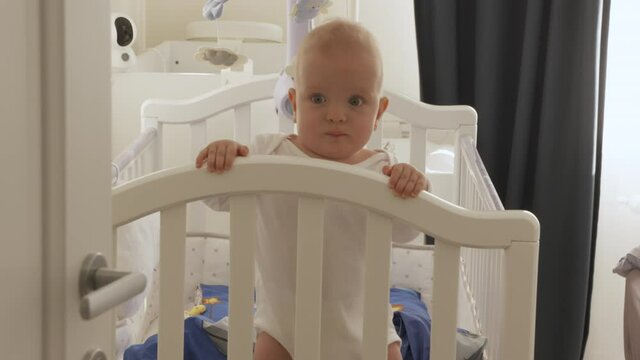 Baby boy standing in white wooden crib holding hands on the rail of rocking cradle, face of cute 7 month old adorable infant closeup. High quality 4k footage