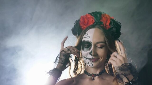 Young dark-haired model with skull makeup wreath on head with choker and in lacy gloves poses in smoke at artificial light in dark studio closeup