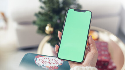 Over the shoulder of young blond woman looking and swiping smartphone with greenscreen and christmas decoration around