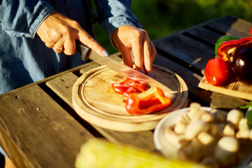 Unrecognizable woman cutting fresh pepper vegetables on wooden board during weekend barbecue in yard, outdoor, prepare for grilling, summer family picnic, food on the nature.