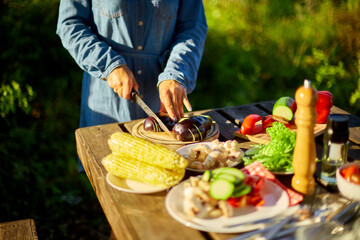 Unrecognizable woman cutting fresh eggplant vegetables on wooden board during weekend barbecue in yard, outdoor, prepare for grilling, summer family picnic, food on the nature.