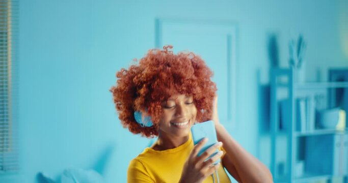 Slow motion portrait of attractive young black woman listening to music on mobile phone application with headphones and dancing in studio. Afro girl enjoying music on smart phone app with headset