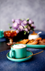 Turquoise cup of coffee with milk foam on the background of a vase with flowers and a plate of cheesecakes