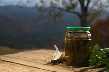 Jar of delicious pickled cucumbers and ingredients on wooden table in mountains, space for text