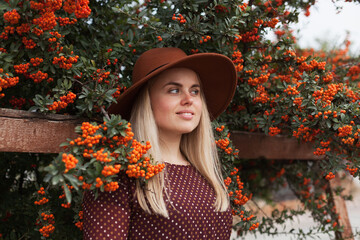 Beautiful woman wearing terracotta brown colous hat and rustic dress near rowan tree with ripe orange berries. Slow living and natural beauty concept.