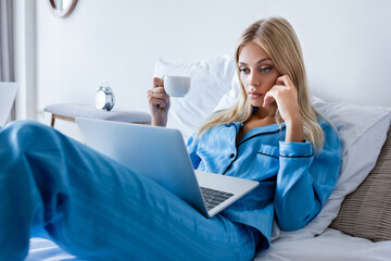 Fototapeta premium young woman in pajamas using laptop and holding cup of coffee