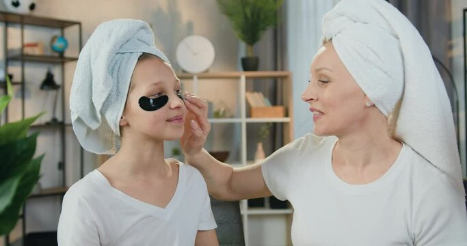 Lovely positive caring loving mother and her teen daughter in terry towels around their heads applying refreshing collagen patches under eyes,skin care concept