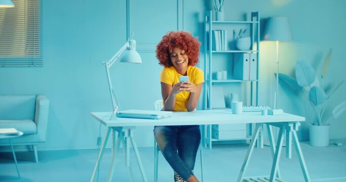 Attractive young black female model with curly hair smiling happy reading message on cellphone while sitting in studio with blue modern interior. Happy cheerful female model using mobile phone at home
