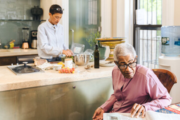 Adult daughter prepares a meal for her mother in their kitchen while elderly mother reads the...