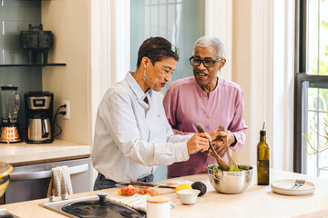 Adult woman prepares a salad while her elderly mother smiles and helps