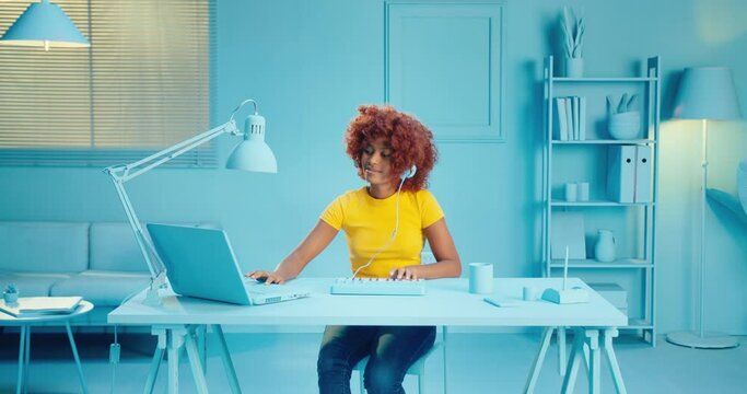 Slow motion of creative black woman with curly hair arranging song using laptop computer and midi keyboard synthesizer in home studio with blue interior. Female model using midi keyboard and laptop