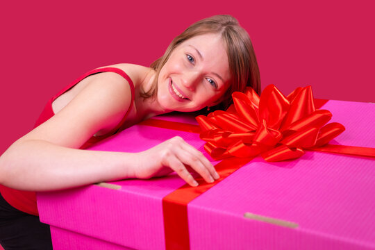 Close up photo of a beautiful happy young woman keeping her head on the big present box of pink color and with bow on that box.