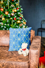 The room is decorated with New Year's decor. A festive tree decorated with New Year's toys next to a brown sofa. On the couch, a blue pillow and a white teddy bear